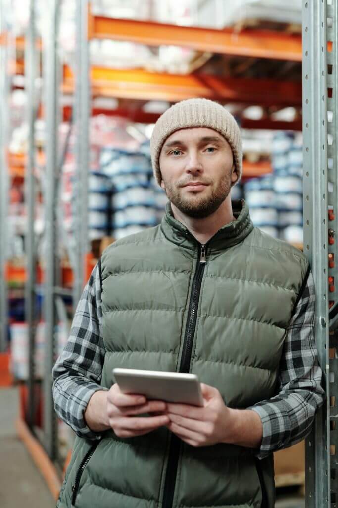 pexels-photo-4484072-4484072 A man wearing a beanie and vest holding a tablet in a warehouse setting.